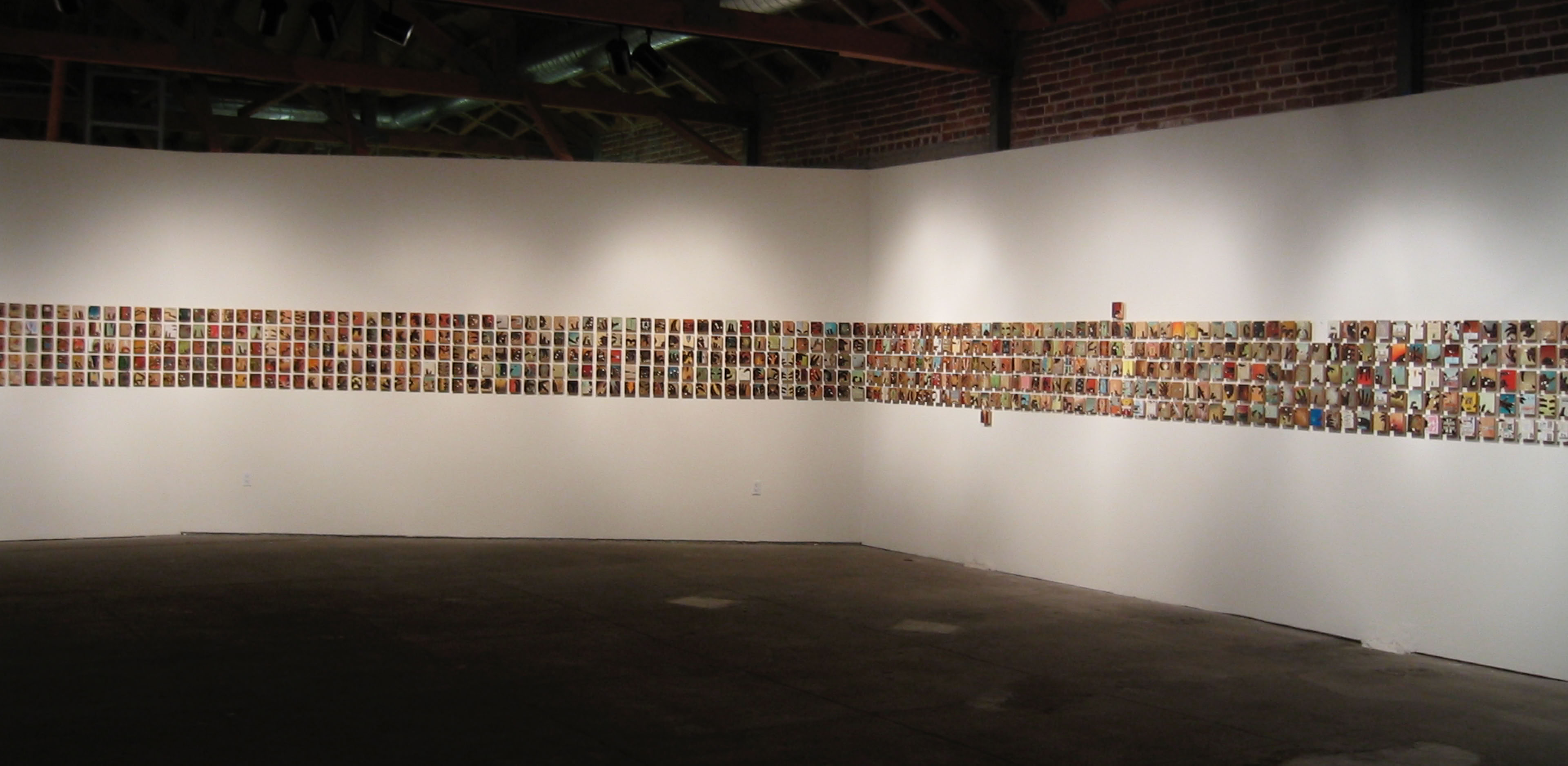 A dimly lit gallery space with a long strip of small, colorful images displayed on a white wall. The ceiling has exposed beams and pipes.